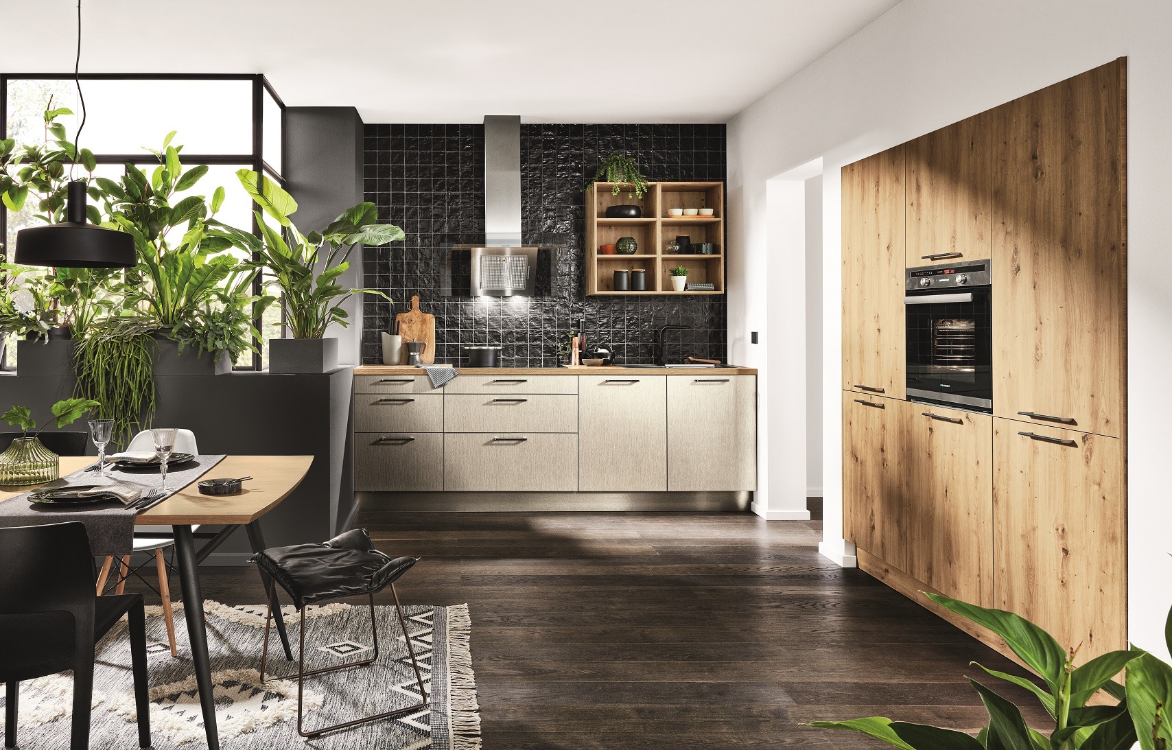 Nowoczesna kuchnia z drewnianą zabudową i czarnymi płytkami na ścianie. Na pierwszym planie stół z nakryciem i rośliny doniczkowe.