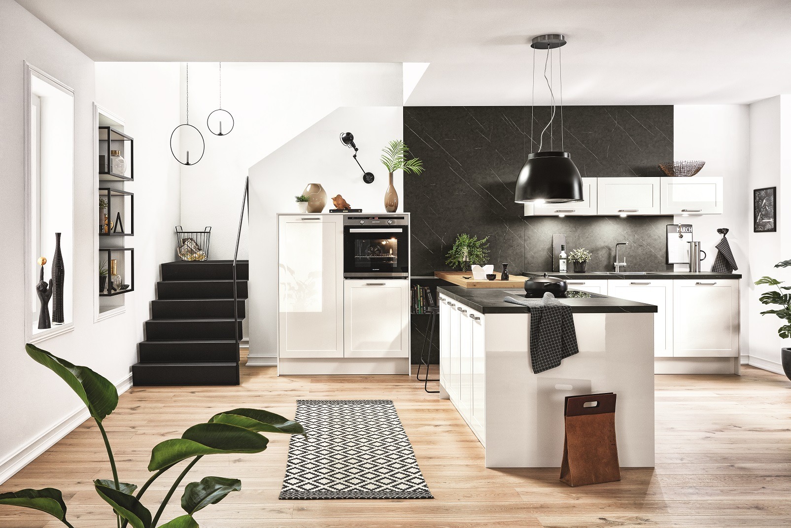 Biała kuchnia z wyspą i czarnym blatem, oświetlona wiszącą lampą. W tle schody i dekoracje. Minimalistyczny dywanik na drewnianej podłodze.