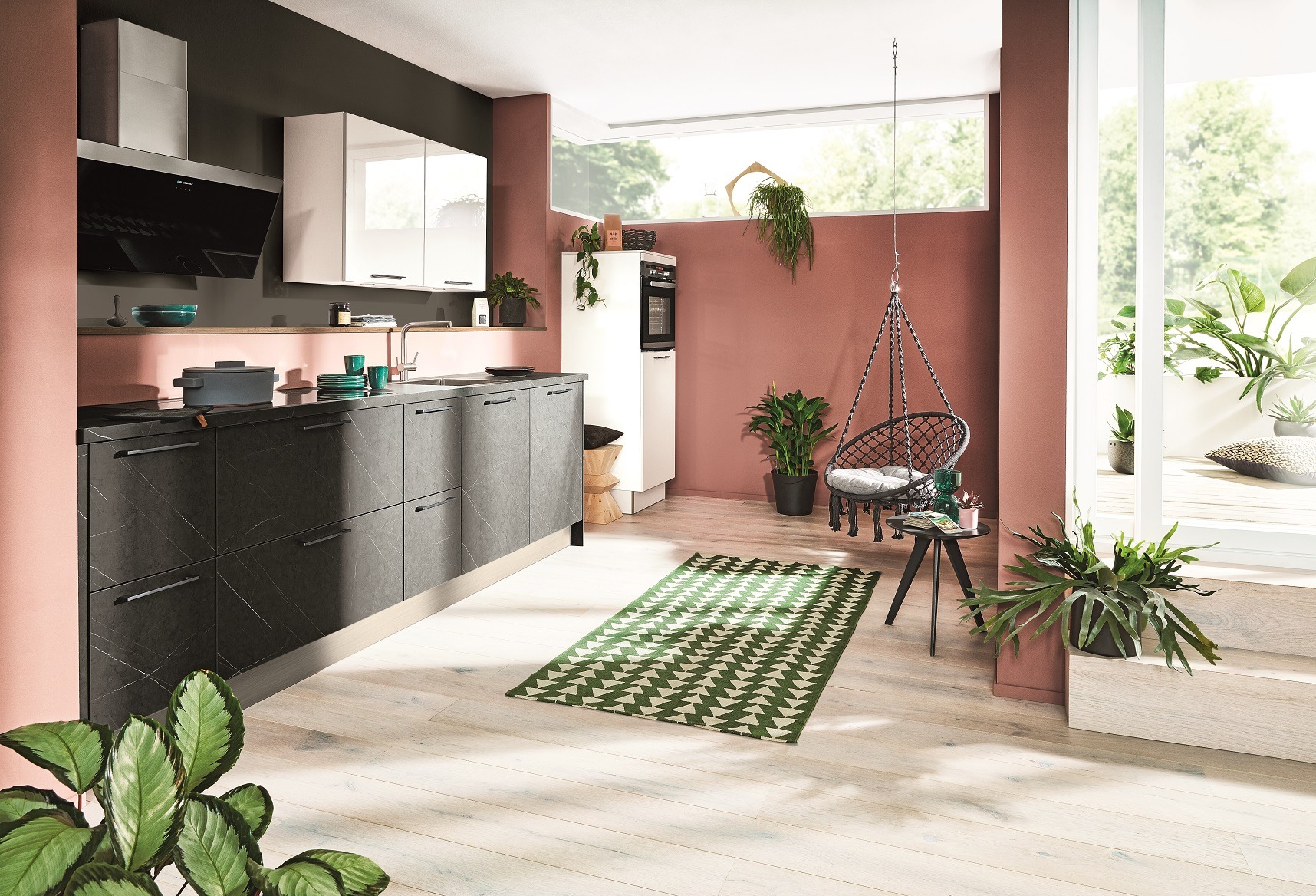 Nowoczesna kuchnia z szarymi szafkami i czarnym blatem, akcentowana roślinami i wiszącym fotelem. Ściany w odcieniach różu i szarości. Jasna podłoga.