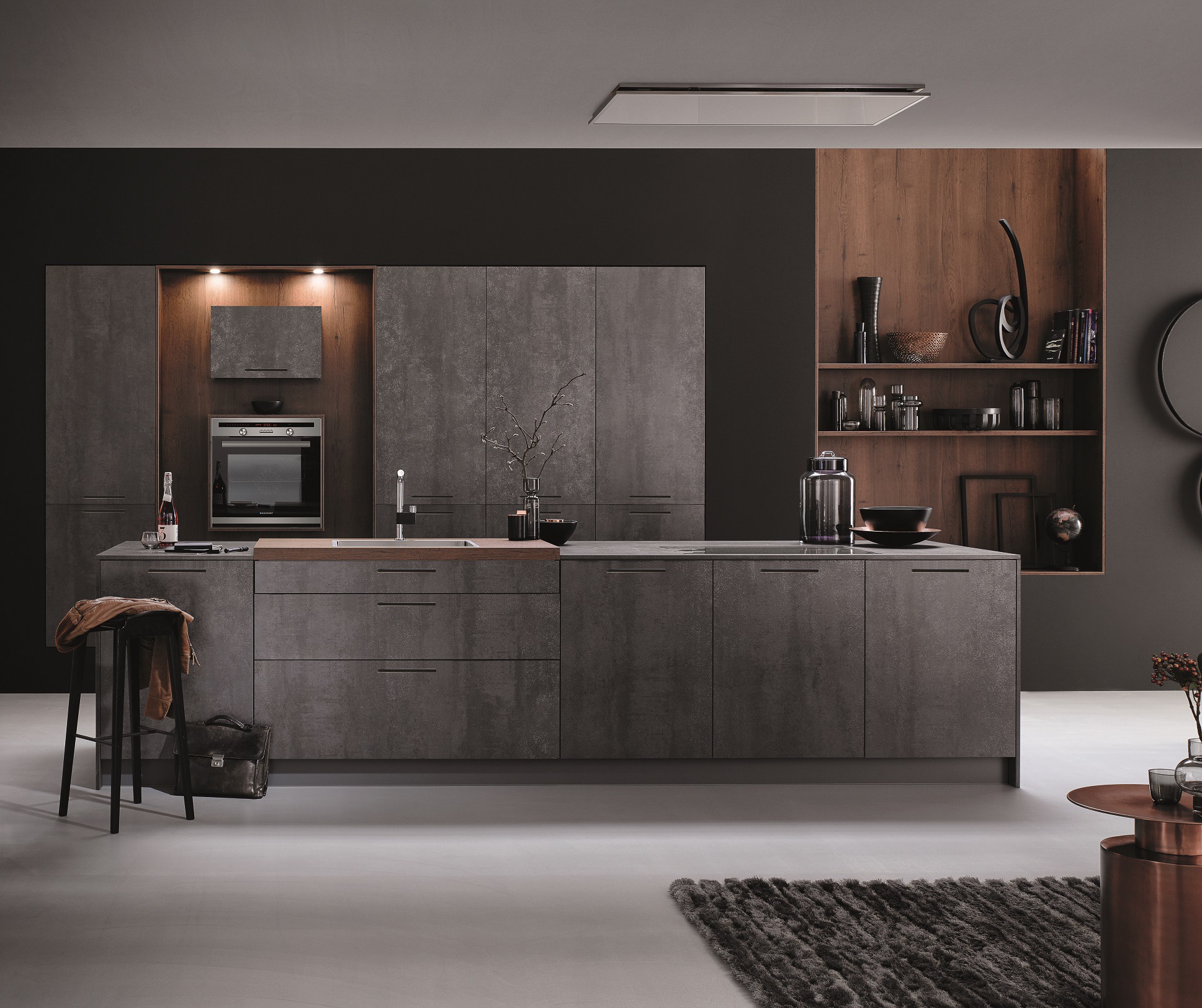 Nowoczesna, grafitowa kuchnia z wyspą i drewnianymi akcentami. Na blacie dekoracje, obok stołek z kurtką i torbą. Minimalistyczny design, oświetlenie punktowe.