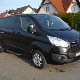 Czarny bus Ford Transit z przyciemnianymi szybami, zaparkowany na brukowanej drodze przed domami jednorodzinnymi w słoneczny dzień.