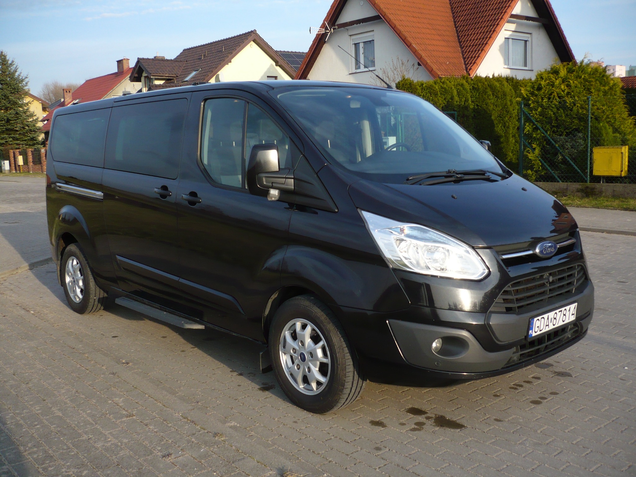 Czarny bus Ford Transit z przyciemnianymi szybami, zaparkowany na brukowanej drodze przed domami jednorodzinnymi w słoneczny dzień.