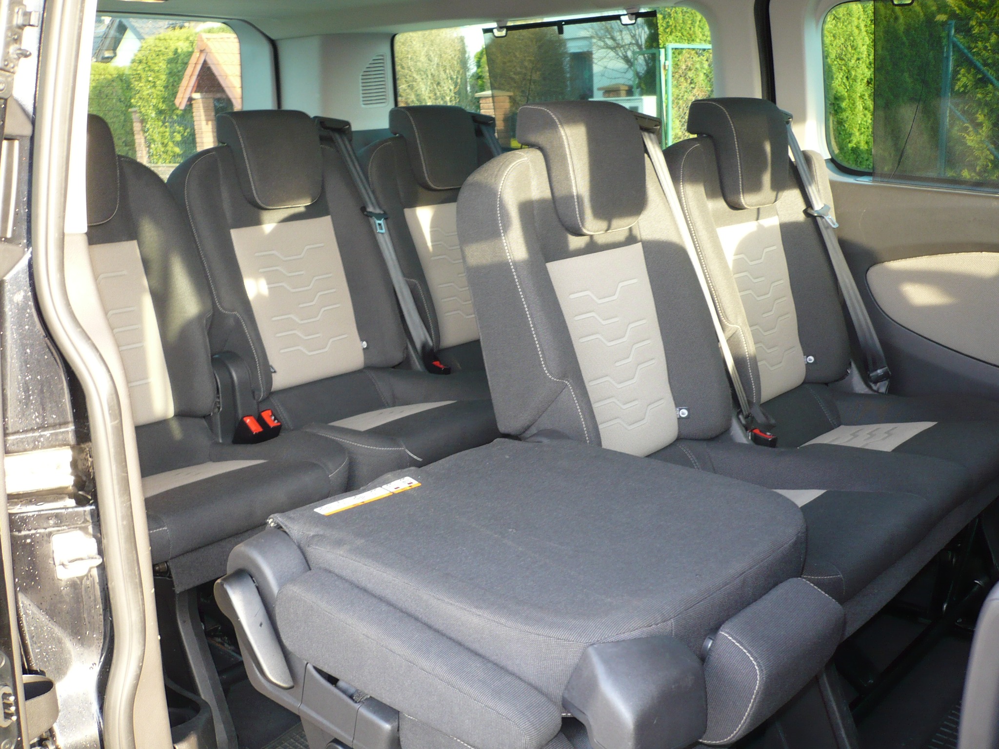 Wnętrze busa pasażerskiego z rozłożonymi, szarymi fotelami z beżowymi wstawkami i widocznymi pasami bezpieczeństwa, widok z otwartych drzwi.