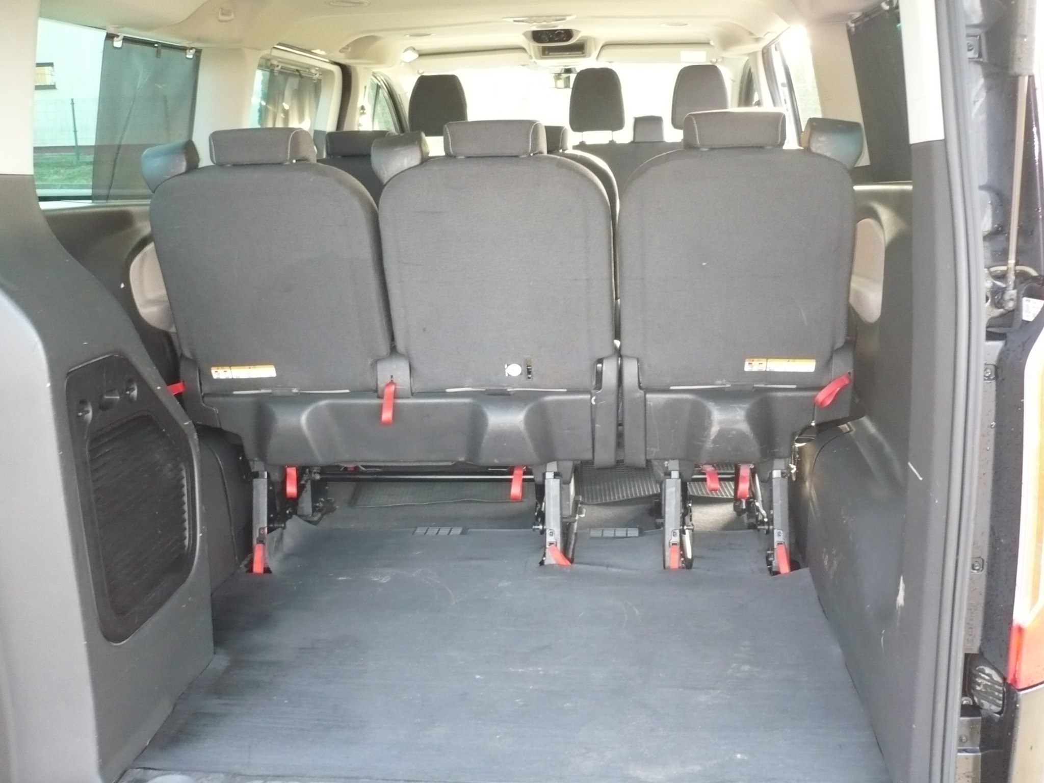Wnętrze busa z trzema rzędami szarych foteli, widok z tyłu, z elementami mocowania foteli i czerwonymi pasami bezpieczeństwa.