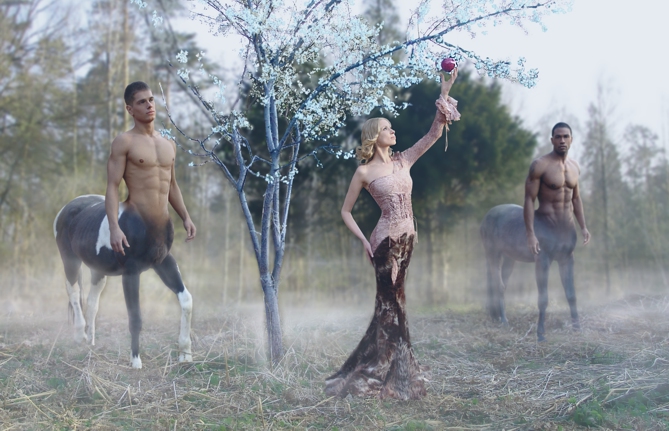Kobieta w ozdobnej sukni sięga po jabłko z drzewa, otoczona dwoma centaurami na tle zalesionego krajobrazu w lekkiej mgle. Scena przypomina baśniową kampanię reklamową.