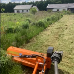 Pomarańczowa kosiarka bijakowa w trakcie pracy na zarośniętej działce, w tle dwa domy jednorodzinne i las.