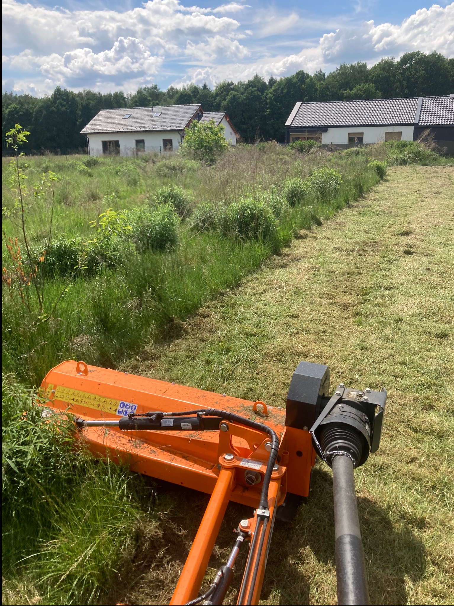 Pomarańczowa kosiarka bijakowa w trakcie pracy na zarośniętej działce, w tle dwa domy jednorodzinne i las.