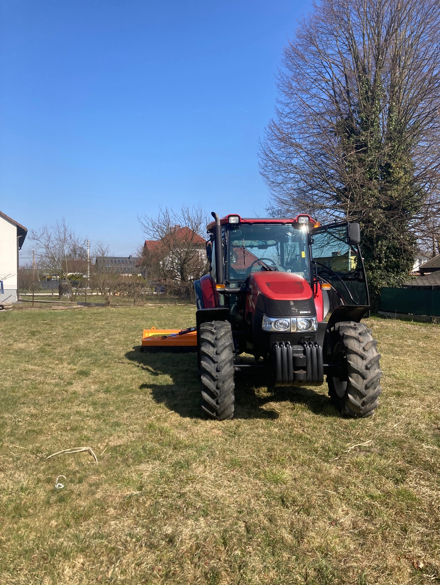 Czerwony traktor z kosiarką na nierównym trawniku w słoneczny dzień. Widoczne budynki w tle i duże drzewo bez liści. Otwarta kabina traktora.