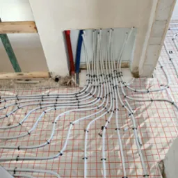 Białe rury systemu ogrzewania podłogowego, mocowane czarnymi uchwytami na folii z czerwoną siatką, wychodzące ze ściany w surowym stanie, z widocznymi fragmentami instalacji wodnej w kolorze...