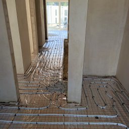 KRIOTTECH - Widok z góry na białe rury ogrzewania podłogowego ułożone na folii z czerwoną siatką w surowym wnętrzu budynku w trakcie budowy, widoczne filary i otwory drzwiowe.