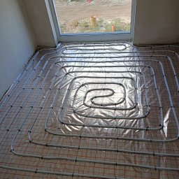 KRIOTTECH - Białe rury instalacji ogrzewania podłogowego ułożone spiralnie na folii z siatką w nowym budynku, widok z góry w kierunku okna dachowego.