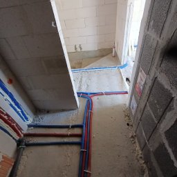 KRIOTTECH - Wnętrze w trakcie prac instalacyjnych: widok na podłogę z poprowadzonymi rurami w kolorach niebieskim i czerwonym, mocowanymi do ściany za pomocą białych opasek. Ściany surowe, z betonu komórkowego...