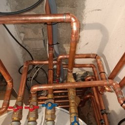 KRIOTTECH - Skrzyżowanie miedzianych rur instalacji wodnej z zaworami i przewodami, widoczne ślady po montażu na surowej ścianie.