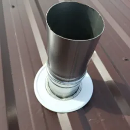 Metalowy komin wentylacyjny wystający z brązowego, blaszane dachu z widocznym uszczelnieniem i datą wykonania zdjęcia w lewym dolnym rogu.