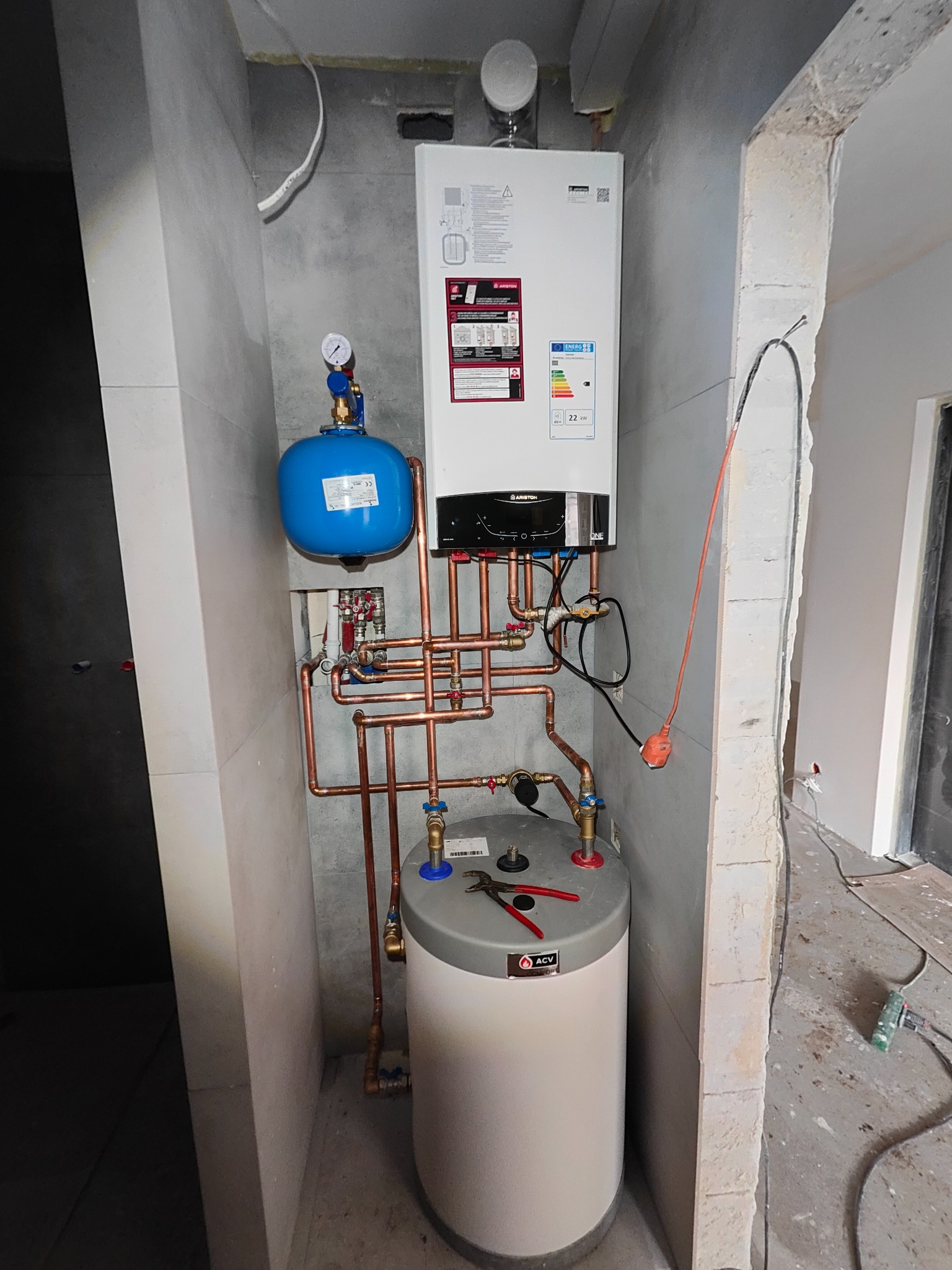 Nowoczesny piec gazowy Ariston zamontowany na szarej ścianie, widoczne miedziane rury, niebieskie naczynie przeponowe i biały zasobnik ACV, czerwone szczypce leżące na zasobniku, surowy stan...