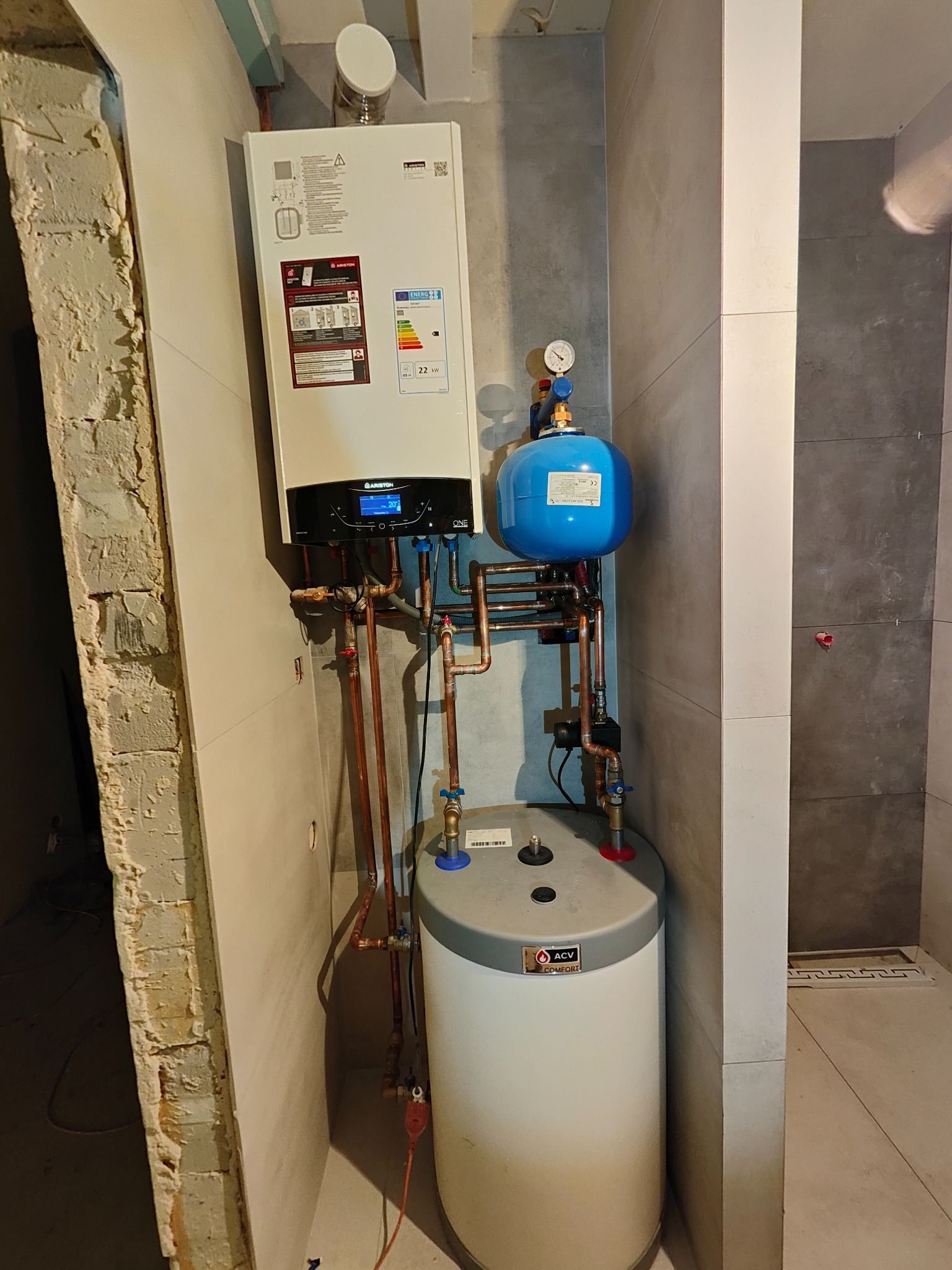 Nowoczesny piec gazowy Ariston ONE z podłączonym zasobnikiem ACV Comfort i niebieskim naczyniem przeponowym, zainstalowany w łazience wykończonej szarymi płytkami, widok na surową ścianę z cegły obok.