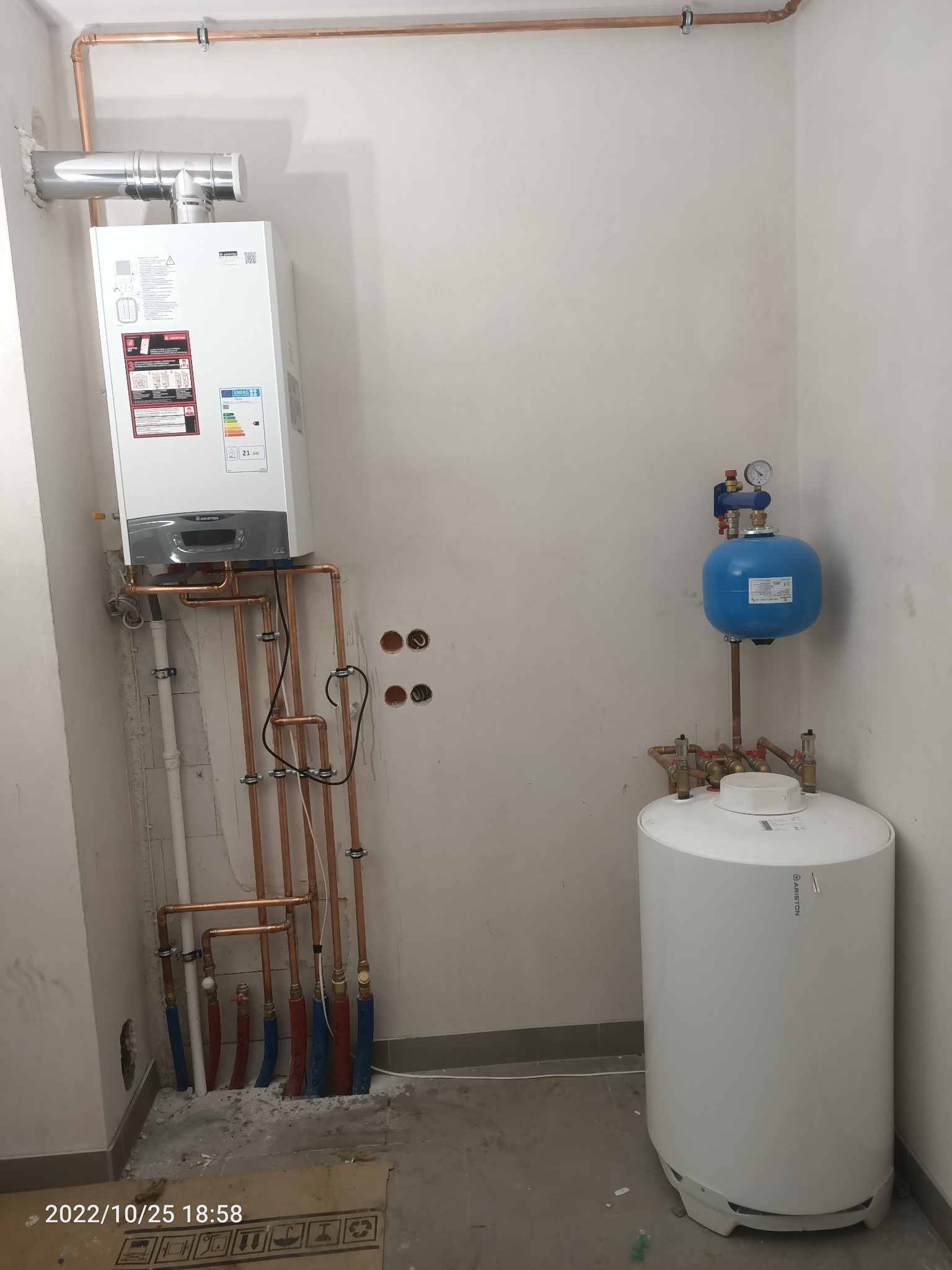 Nowo zainstalowany, wiszący piec gazowy Ariston z podłączonymi miedzianymi rurami, zbiornik wyrównawczy i zasobnik ciepłej wody Ariston w surowym pomieszczeniu z widocznymi śladami prac...