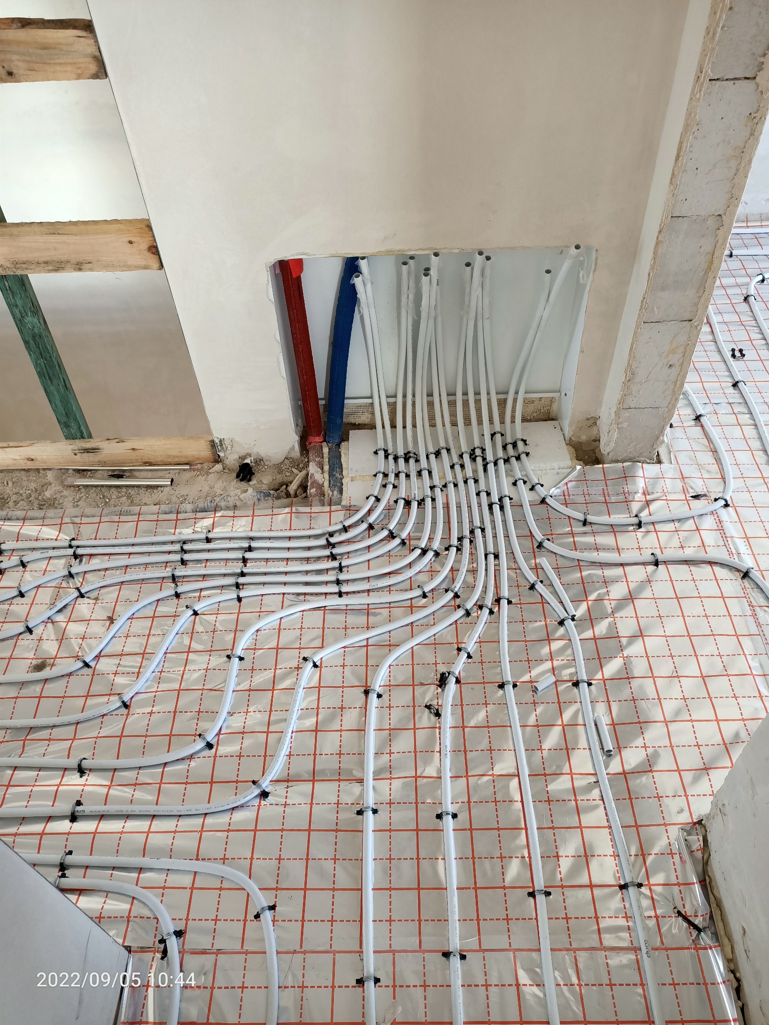 Białe rury systemu ogrzewania podłogowego, mocowane czarnymi uchwytami na folii z czerwoną siatką, wychodzące ze ściany w surowym stanie, z widocznymi fragmentami instalacji wodnej w kolorze...