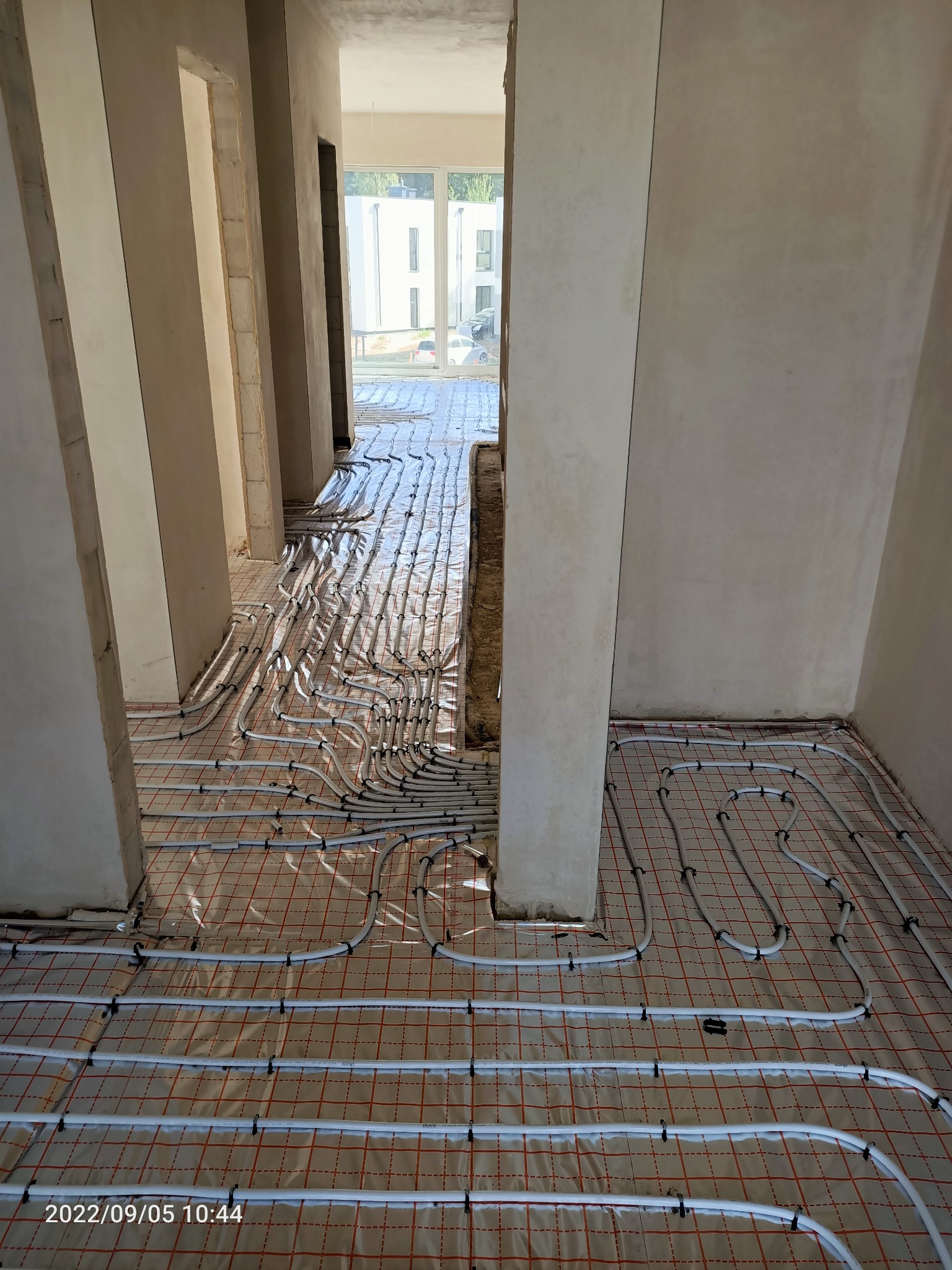 Widok z góry na białe rury ogrzewania podłogowego ułożone na folii z czerwoną siatką w surowym wnętrzu budynku w trakcie budowy, widoczne filary i otwory drzwiowe.