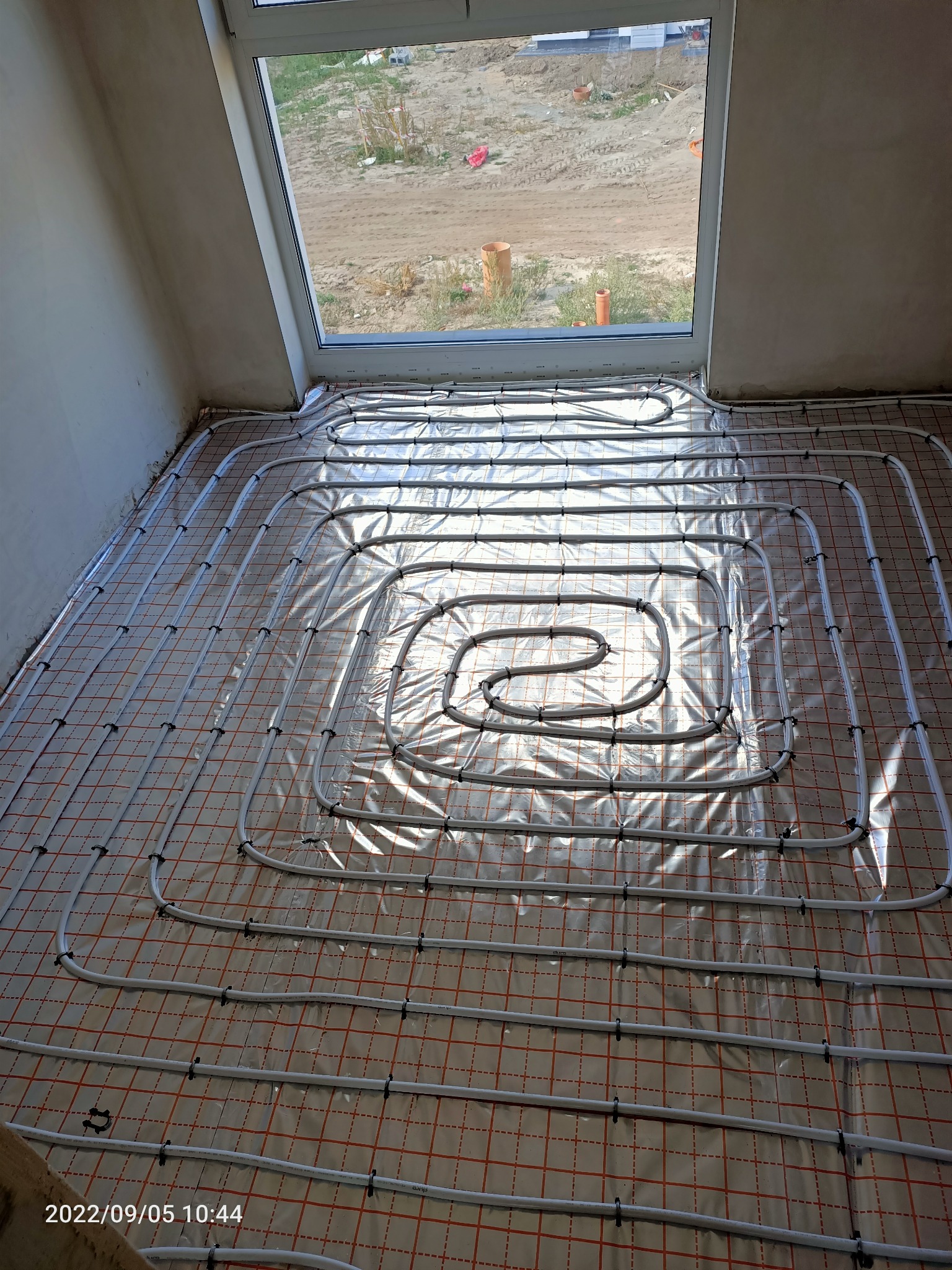 Białe rury instalacji ogrzewania podłogowego ułożone spiralnie na folii z siatką w nowym budynku, widok z góry w kierunku okna dachowego.