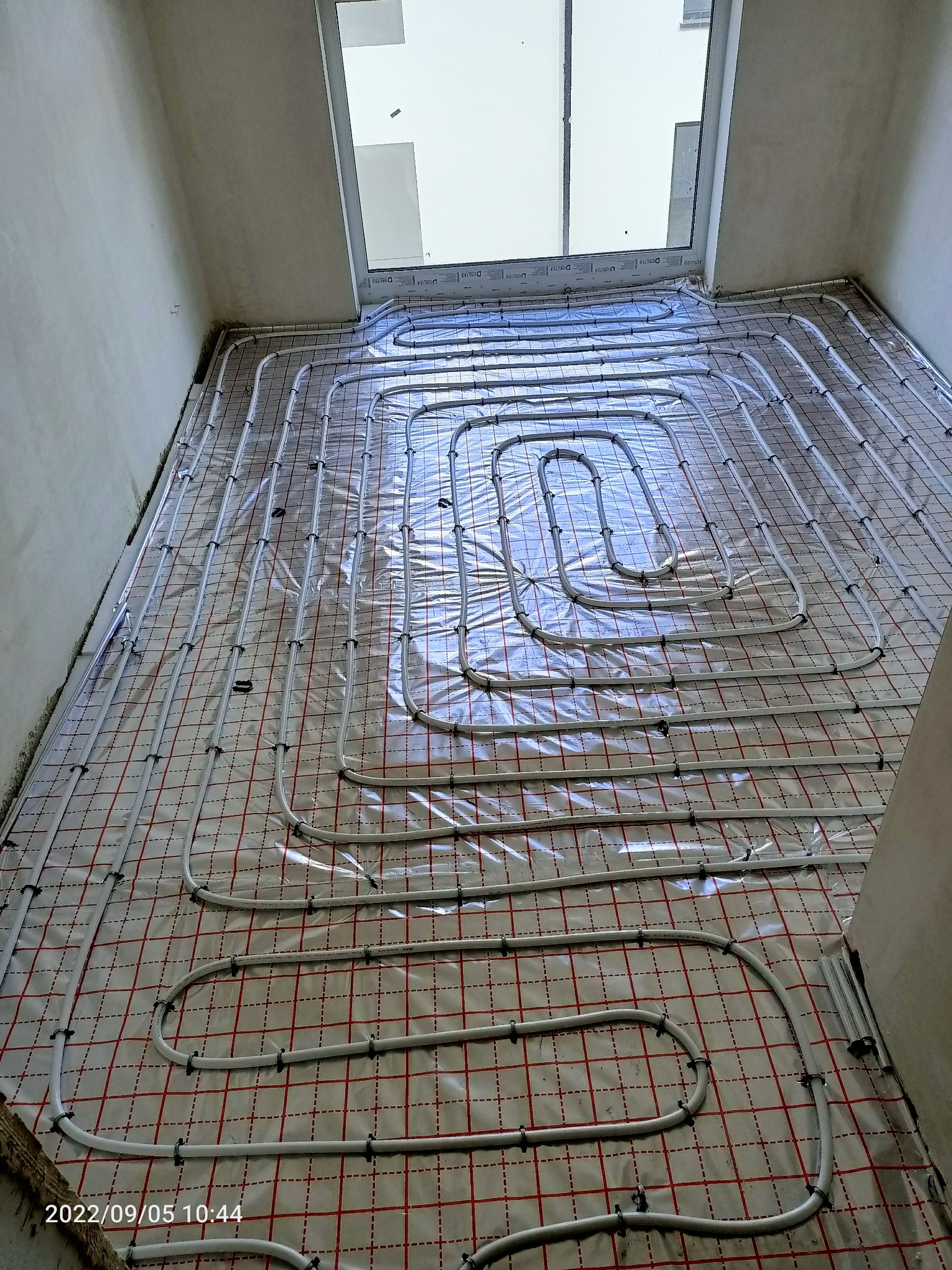 Białe rury systemu ogrzewania podłogowego, ułożone na folii z czerwoną siatką, w surowym pomieszczeniu z oknem w górnej części ściany.