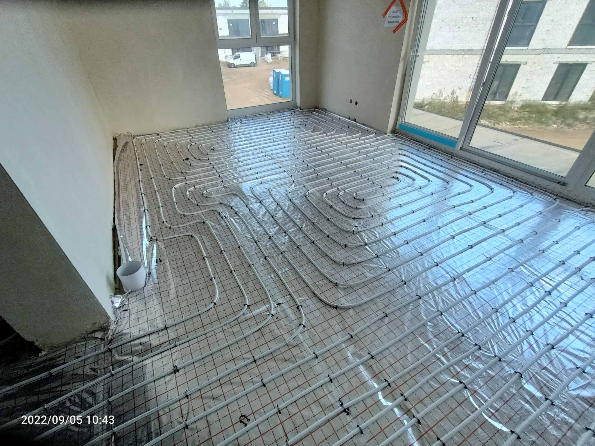 Rozłożona instalacja ogrzewania podłogowego na folii z siatką, widok z góry, surowy stan wykończenia wnętrza, widoczne okna i częściowo budynek na zewnątrz.