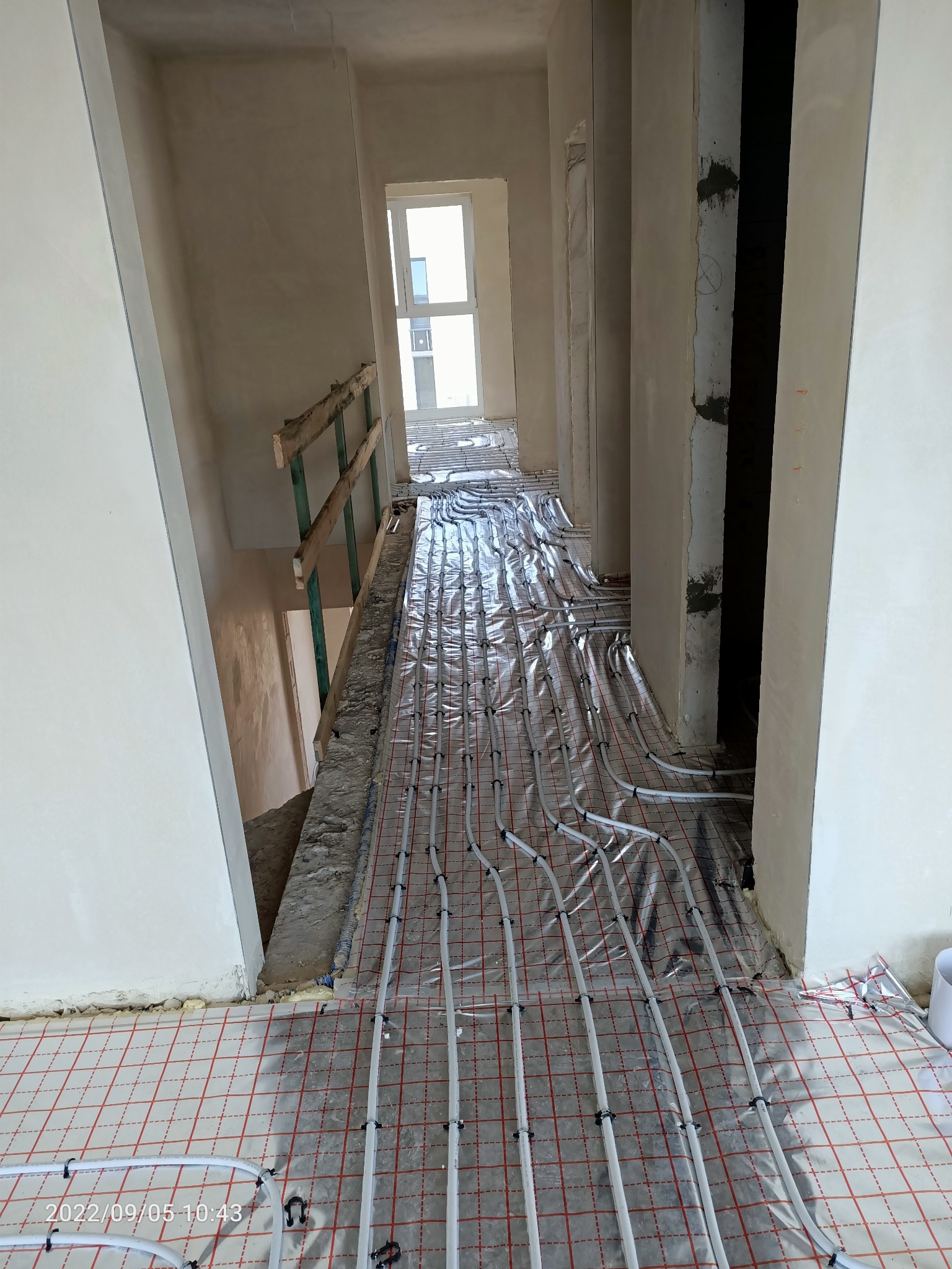 Białe rury ogrzewania podłogowego rozłożone na folii z czerwoną siatką w surowym wnętrzu domu w trakcie budowy, widok z góry na korytarz z oknem w oddali.