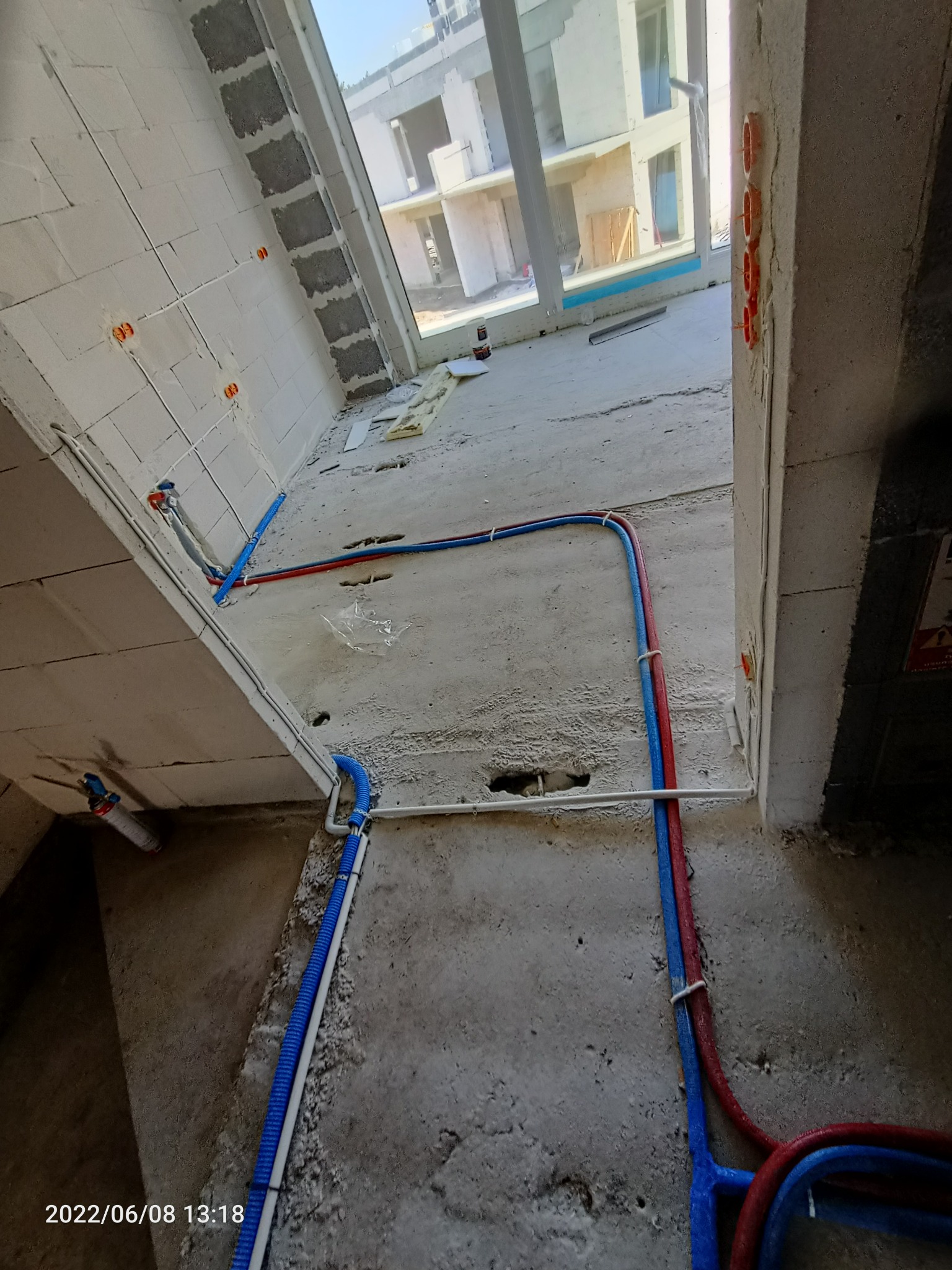 Wnętrze w trakcie prac instalacyjnych: widoczne poprowadzone rury instalacji wodnej (czerwone i niebieskie) przy ścianie z betonu komórkowego oraz peszel instalacji elektrycznej, okrągłe puszki...