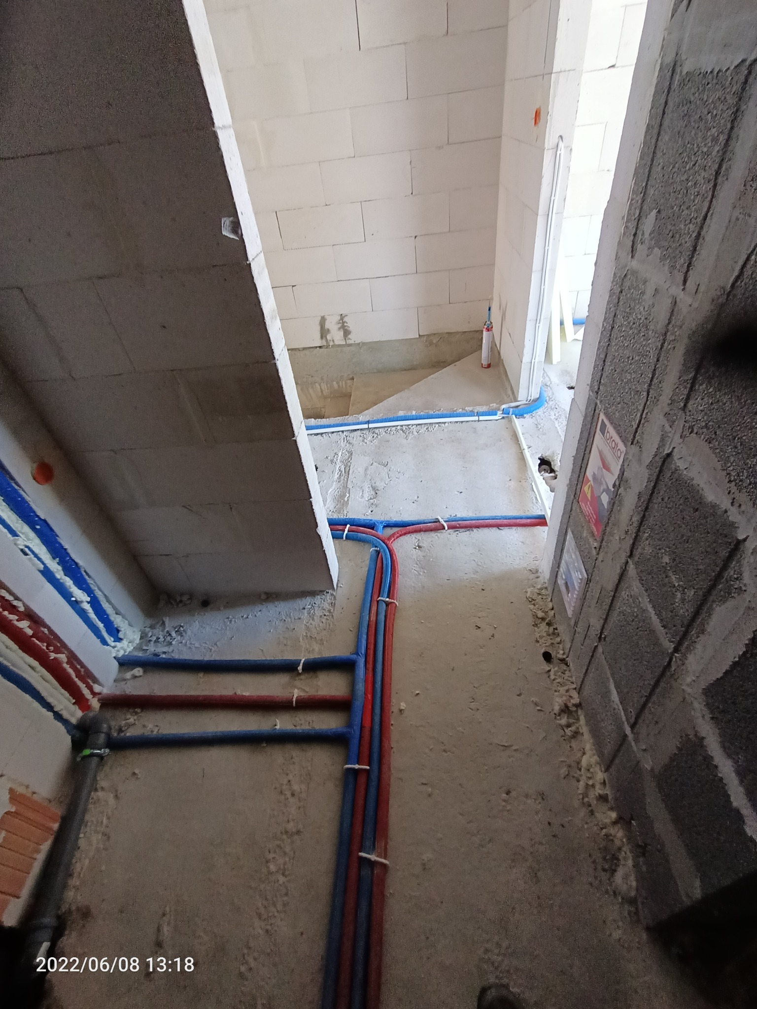 Wnętrze w trakcie prac instalacyjnych: widok na podłogę z poprowadzonymi rurami w kolorach niebieskim i czerwonym, mocowanymi do ściany za pomocą białych opasek. Ściany surowe, z betonu komórkowego...
