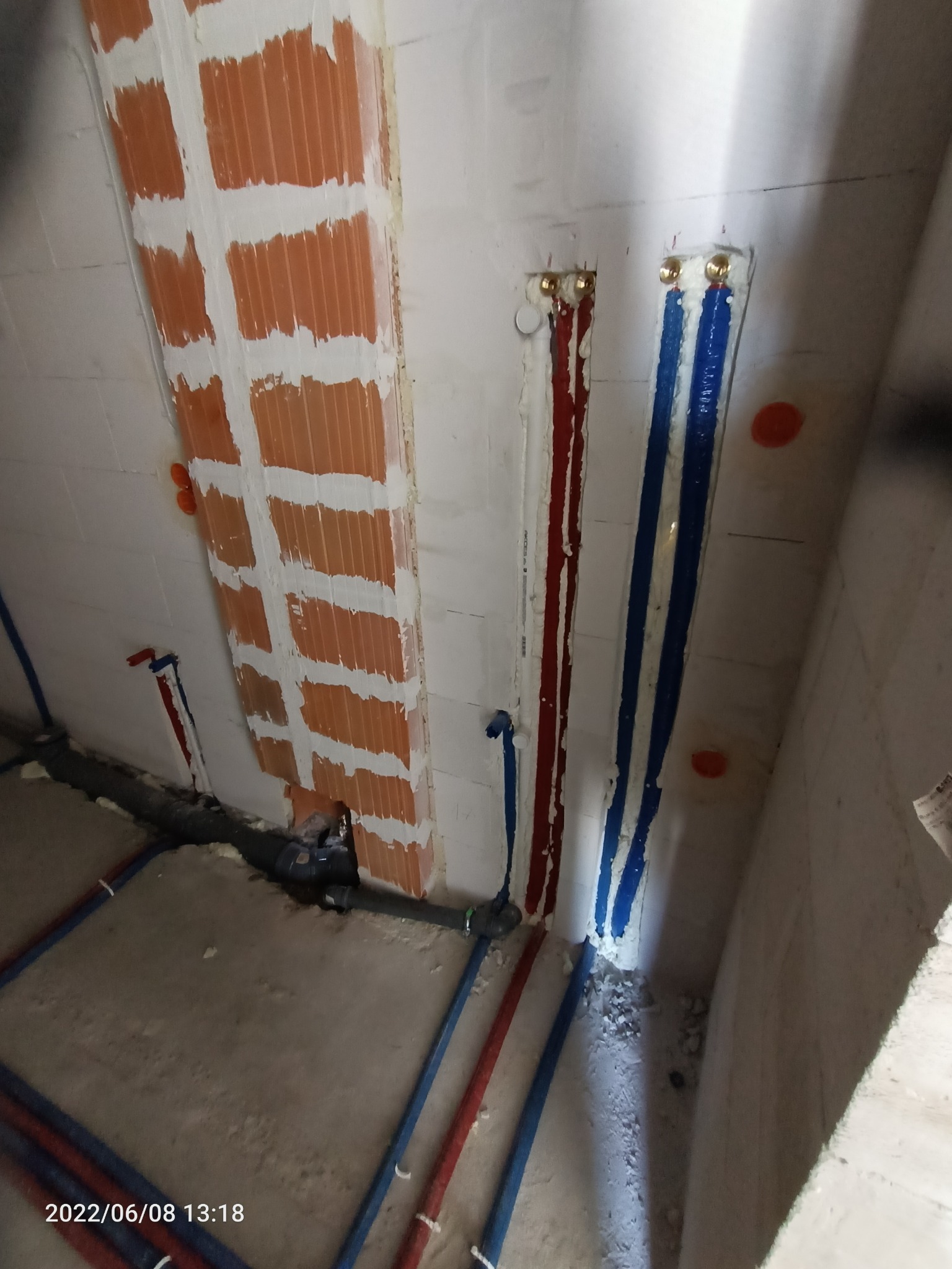 Świeżo położone rury hydrauliczne w kolorach czerwonym i niebieskim, prowadzone wzdłuż ścian, z widocznymi fragmentami muru i zaprawy. Część instalacji ukryta w bruzdach w ścianie.