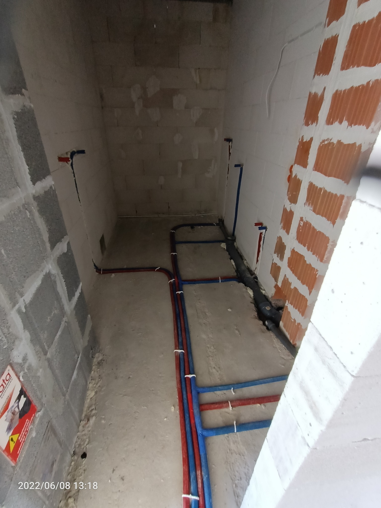 Surowy stan łazienki w trakcie montażu instalacji wodno-kanalizacyjnej, widoczne rury w kolorze niebieskim i czerwonym, przytwierdzone do podłoża za pomocą białych uchwytów, ściany z bloczków...