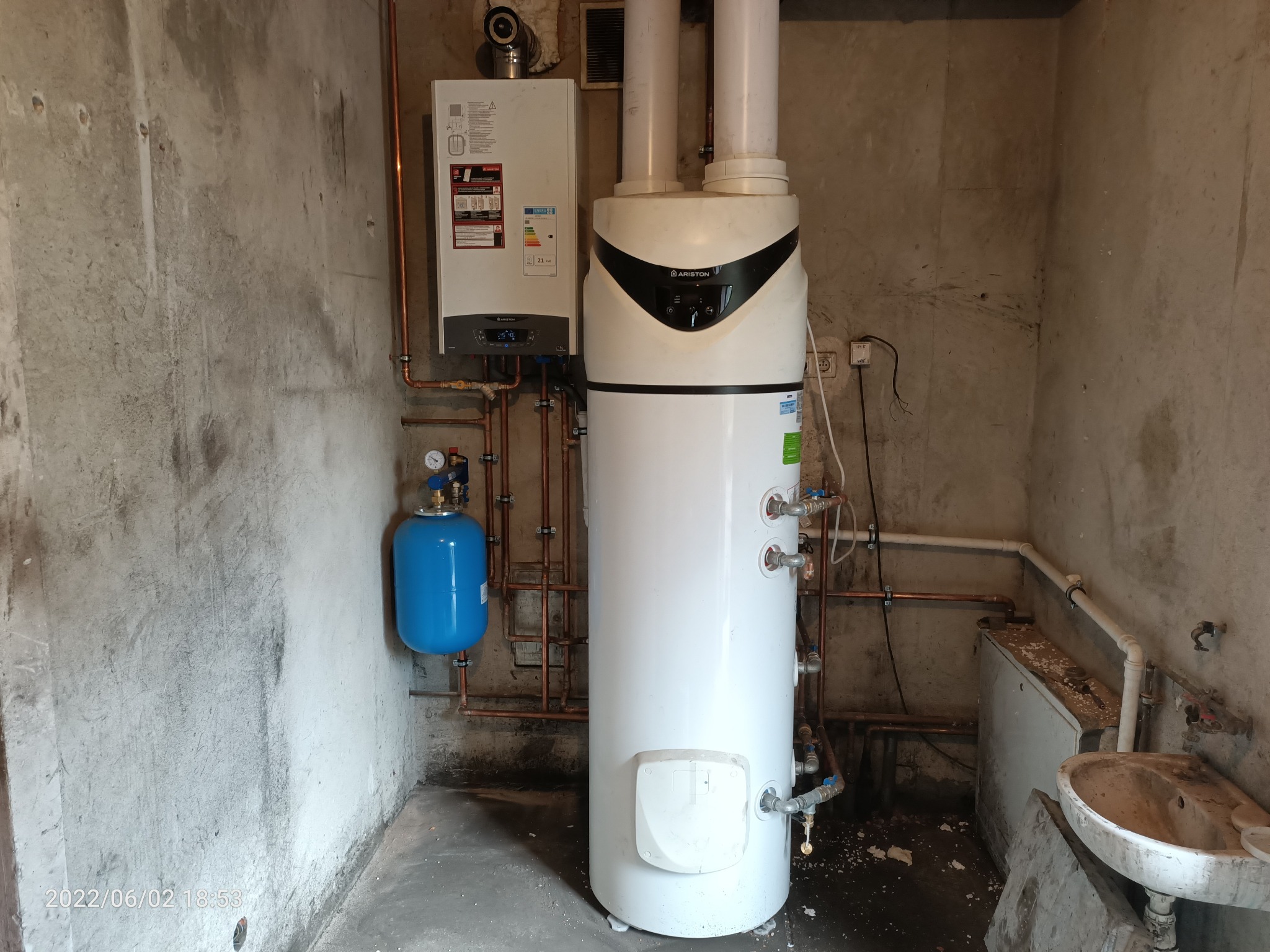 Nowoczesny piec gazowy Ariston z zasobnikiem, podłączony miedzianymi rurami, z niebieskim zbiornikiem wyrównawczym na tle surowej, betonowej ściany w kotłowni.