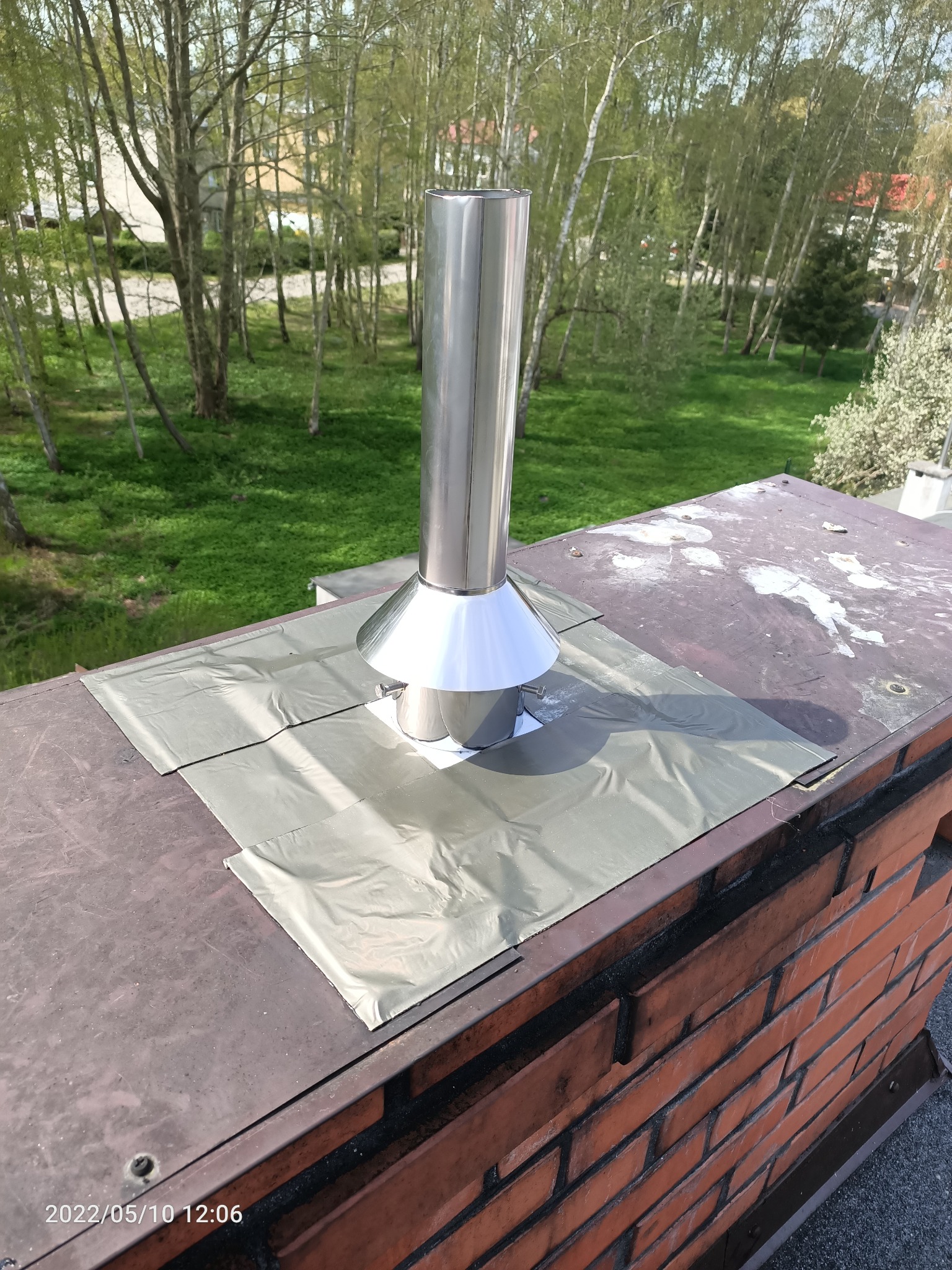 Nowy, błyszczący, metalowy komin zamontowany na ceglanym kominie dachowym, zabezpieczony srebrną folią uszczelniającą, z widokiem na zielone drzewa w tle.