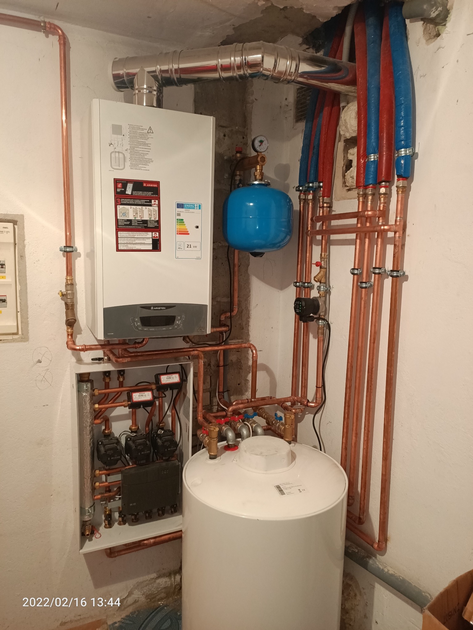Biały piec gazowy Ariston zamontowany na ścianie, z licznymi miedzianymi rurami, niebieskim zbiornikiem wyrównawczym i białym zasobnikiem ciepłej wody, widoczne również pompy obiegowe w szafce...
