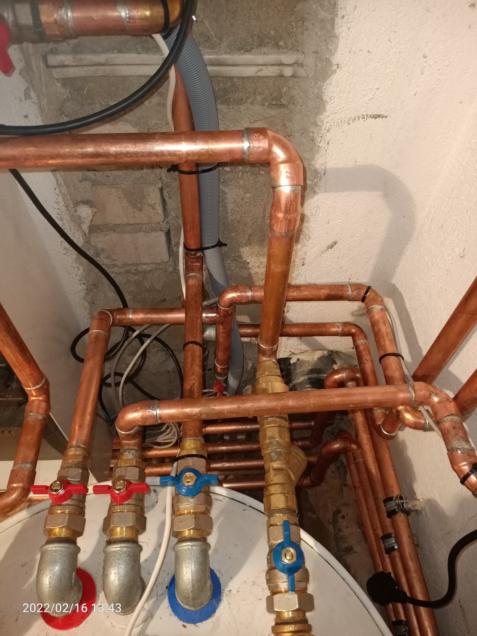 Skrzyżowanie miedzianych rur instalacji wodnej z zaworami i przewodami, widoczne ślady po montażu na surowej ścianie.