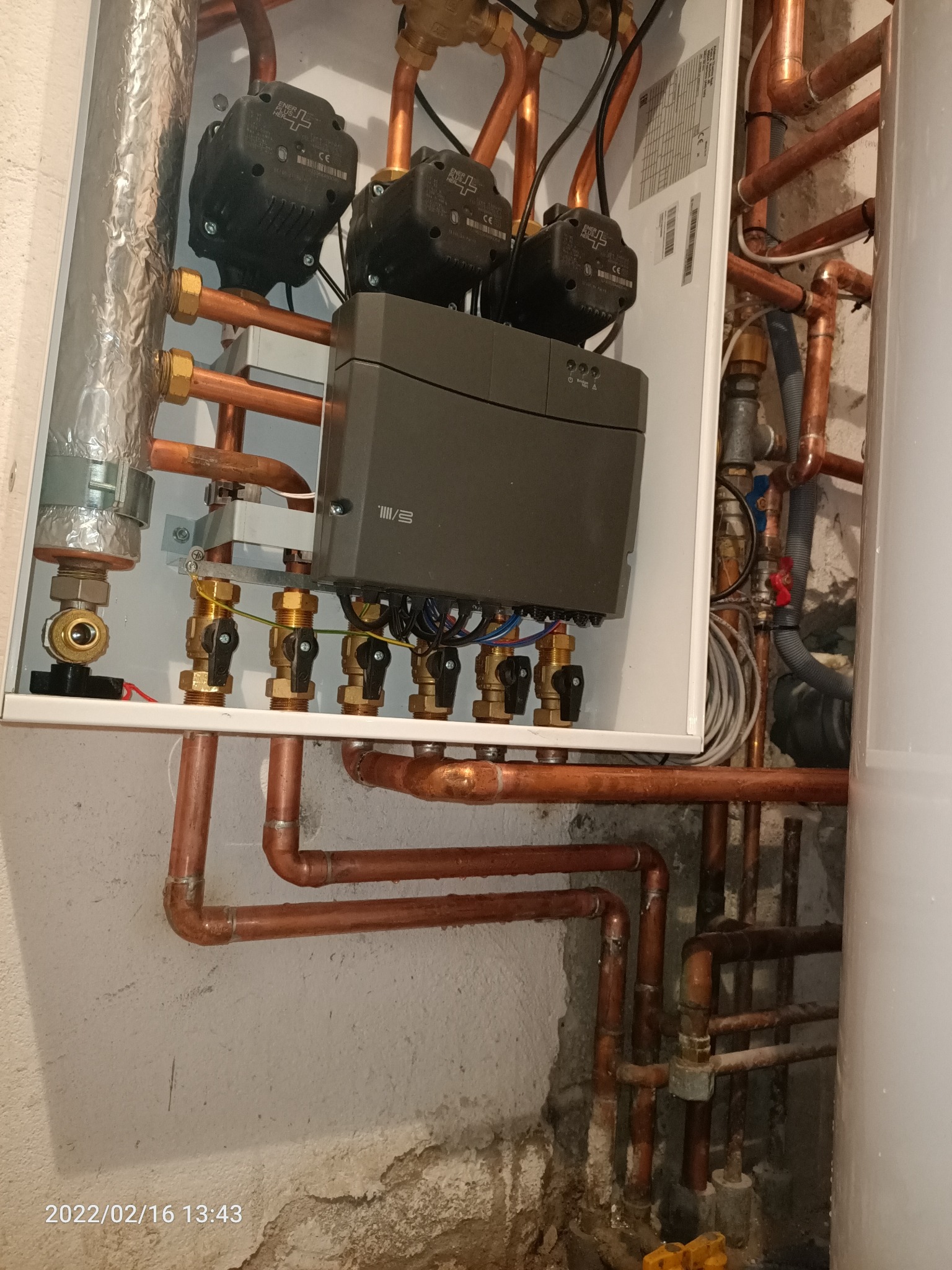Wnętrze szafki z rozdzielaczem ogrzewania podłogowego, widoczne miedziane rury, zawory z czarnymi pokrętłami oraz elektroniczny moduł sterujący. Na zewnątrz szafki widoczny fragment bojlera...