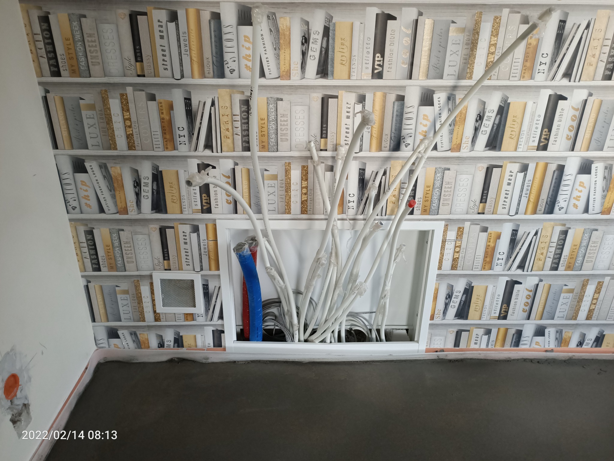 Instalacja elektryczna w trakcie montażu, widoczne białe rurki peszel i kable wystające ze ściany na tle tapety imitującej półki z książkami.