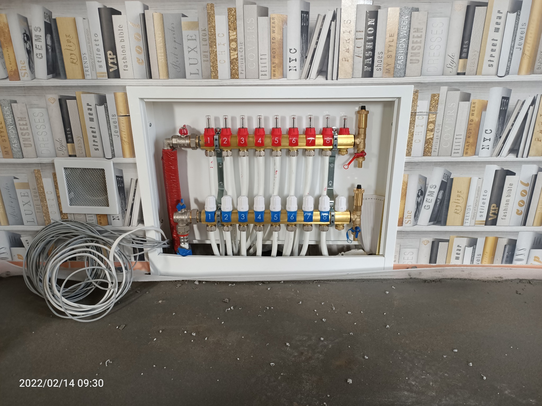 Rozdzielacz ogrzewania podłogowego w szafce, z rurami oznaczonymi numerami od 1 do 8, na tle tapety imitującej regał z książkami.