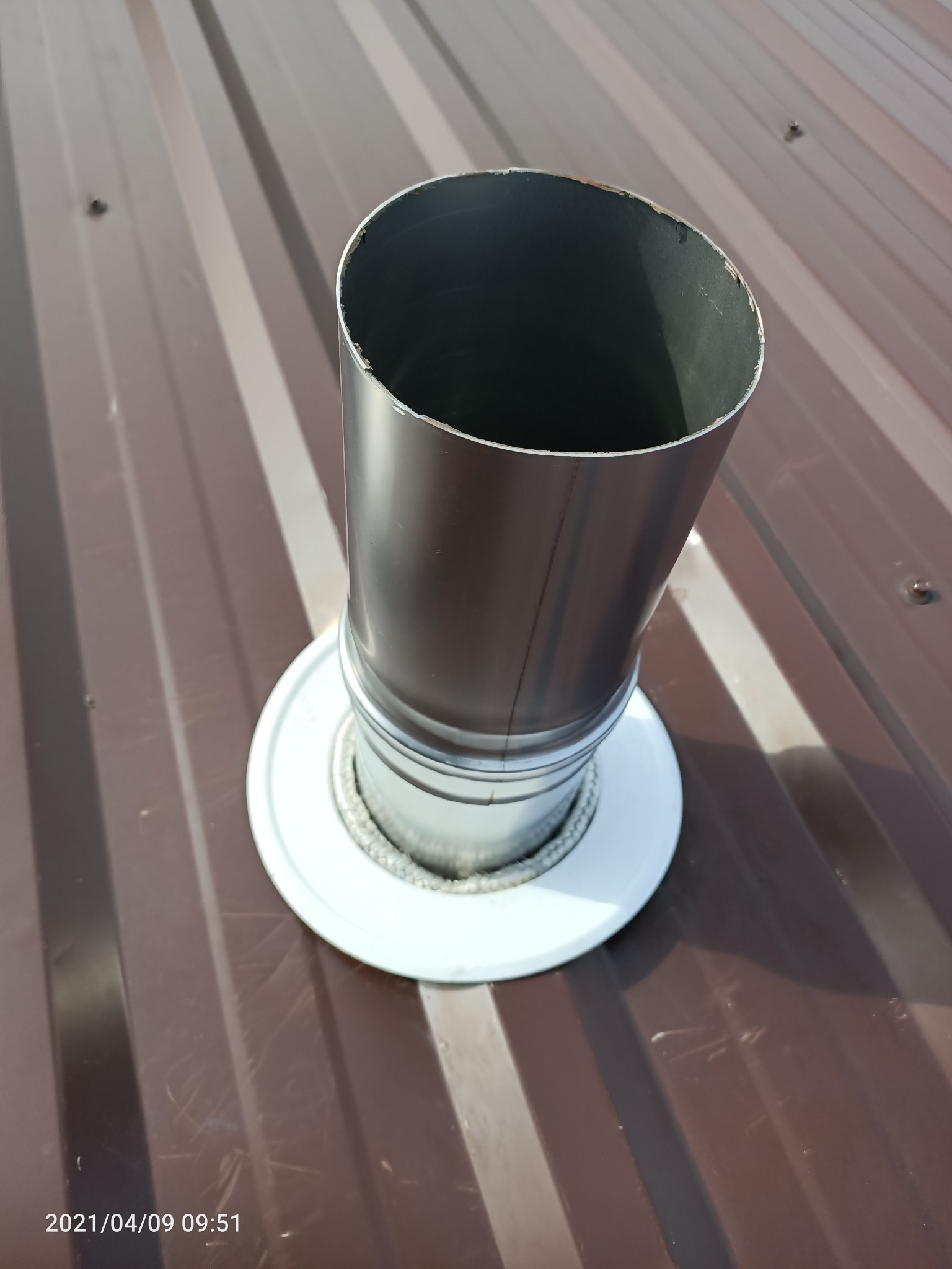 Metalowy komin wentylacyjny wystający z brązowego, blaszane dachu z widocznym uszczelnieniem i datą wykonania zdjęcia w lewym dolnym rogu.