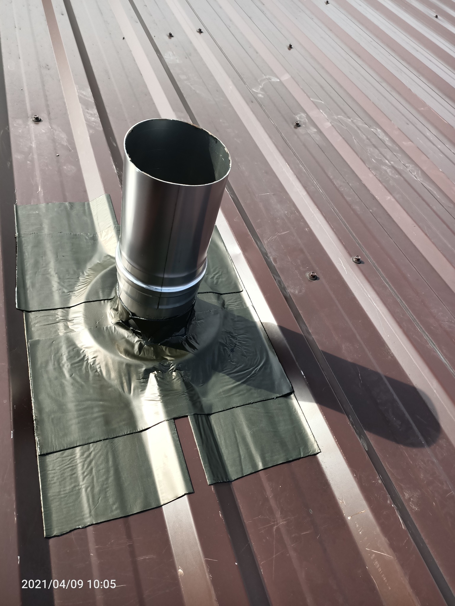 Metalowy komin wentylacyjny wystający z brązowego dachu z blachy trapezowej, uszczelniony czarną taśmą dekarską, widoczny cień.