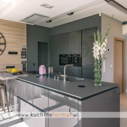 AGNIESZKA ĆWIERTNIAK-JANIA FORMAT HOME - Nowoczesna kuchnia z szarymi, lakierowanymi frontami, wyspą z hokerami, drewnianą ścianą z zegarem i dekoracjami oraz widokiem na korytarz z mapą świata na ścianie.