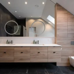 Nowoczesna łazienka na poddaszu z drewnianą zabudową umywalki z szufladami, okrągłymi lustrami z podświetleniem LED i wiszącą toaletą.