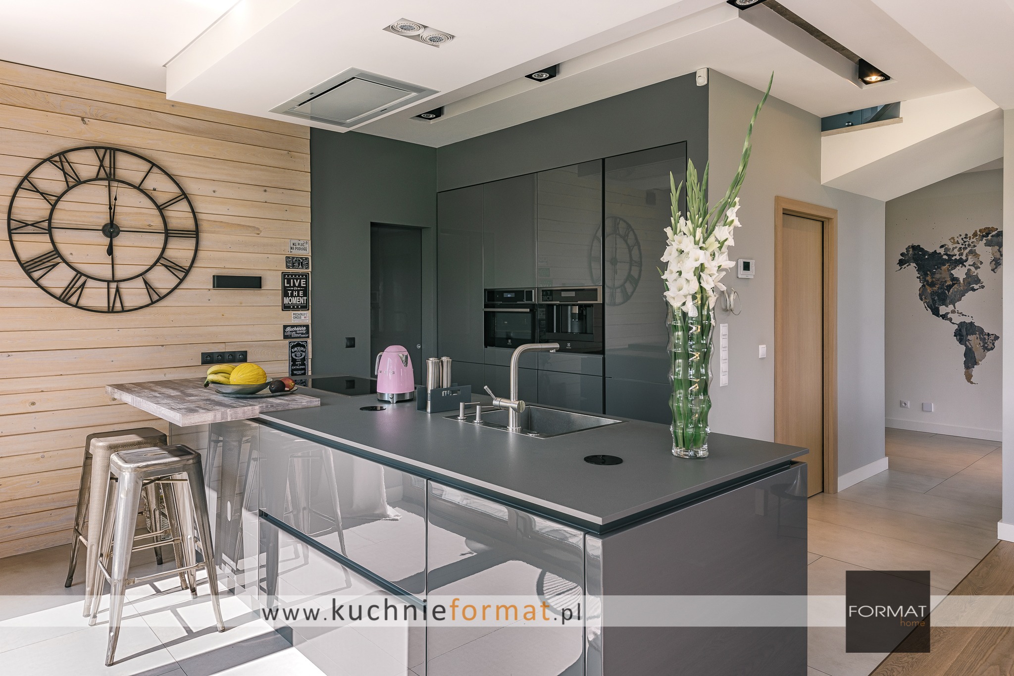 Nowoczesna kuchnia z szarymi, lakierowanymi frontami, wyspą z hokerami, drewnianą ścianą z zegarem i dekoracjami oraz widokiem na korytarz z mapą świata na ścianie.