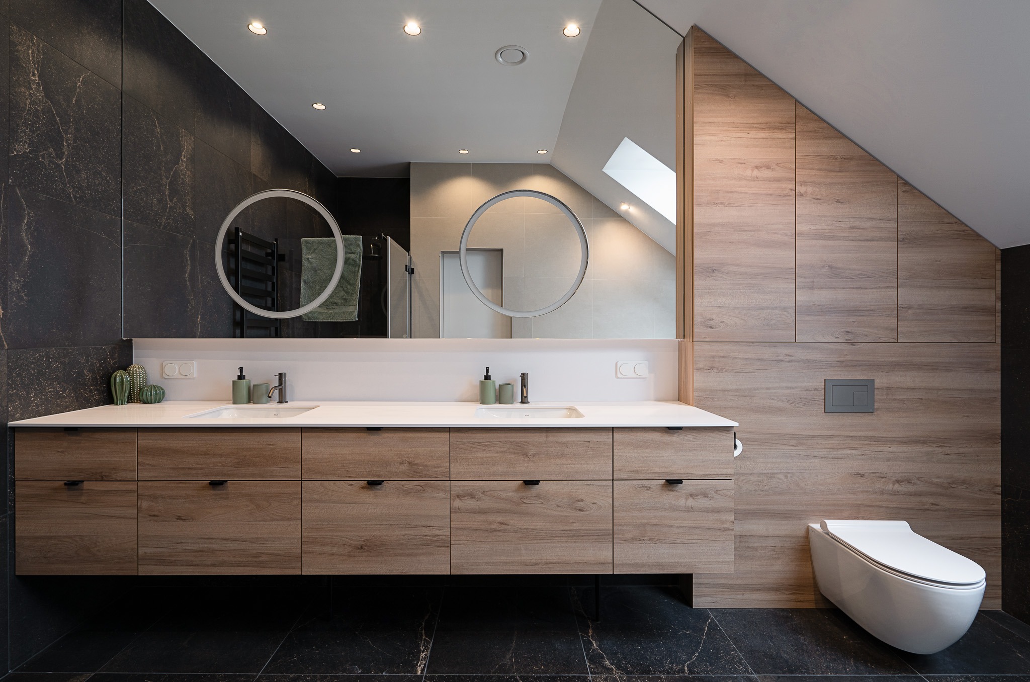 Nowoczesna łazienka na poddaszu z drewnianą zabudową umywalki z szufladami, okrągłymi lustrami z podświetleniem LED i wiszącą toaletą.
