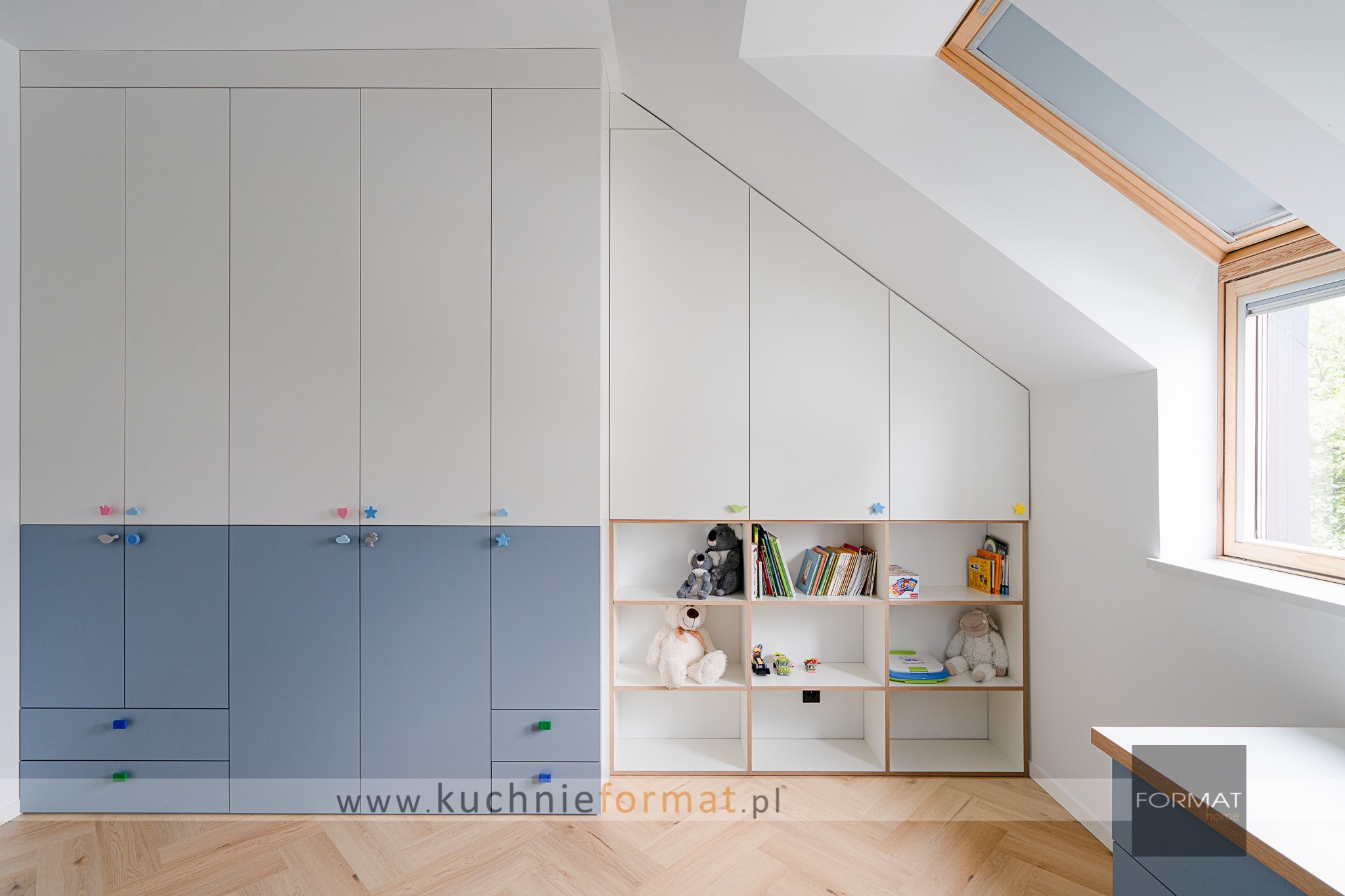 Biało-szara zabudowa meblowa w pokoju dziecięcym na poddaszu ze skośnym sufitem, z szafkami z kolorowymi uchwytami i otwartymi półkami z książkami i zabawkami, parkiet w jodełkę.