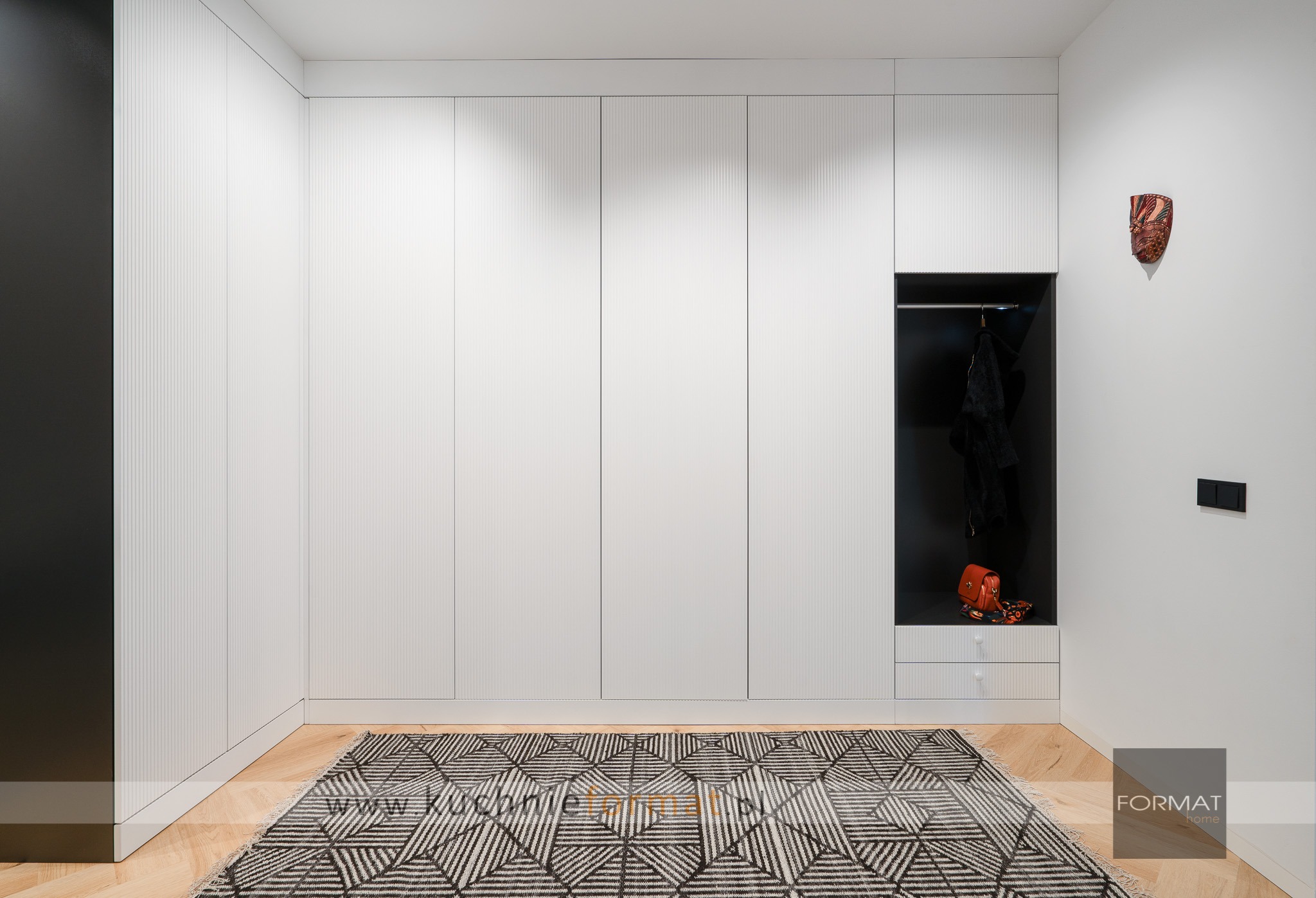 Biała zabudowa szaf w przedpokoju z ryflowanymi frontami, otwartą wnęką z wieszakiem i szufladami, wzorzysty dywan na parkiecie, maska na ścianie.