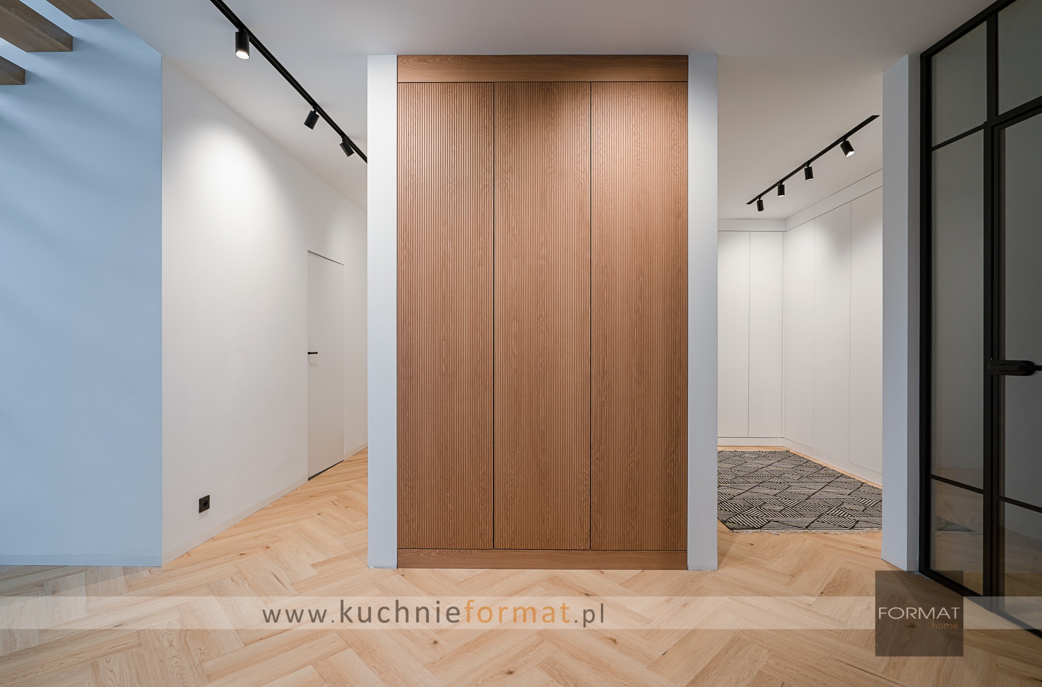 Wnętrze korytarza z drewnianą zabudową szafy z ryflowanymi frontami, oświetlenie szynowe, parkiet w jodełkę, widok na garderobę z dywanem geometrycznym.