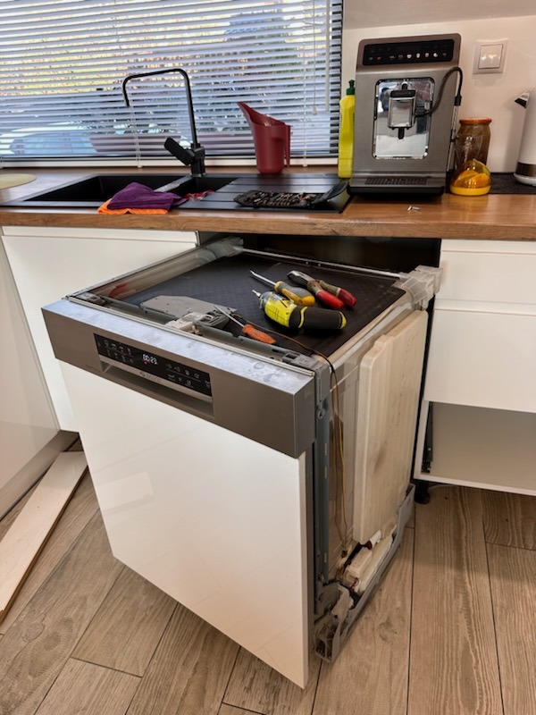 Otwarta zmywarka w trakcie naprawy, widoczne narzędzia na górnej krawędzi, kuchnia w tle z ekspresem do kawy i zlewozmywakiem.