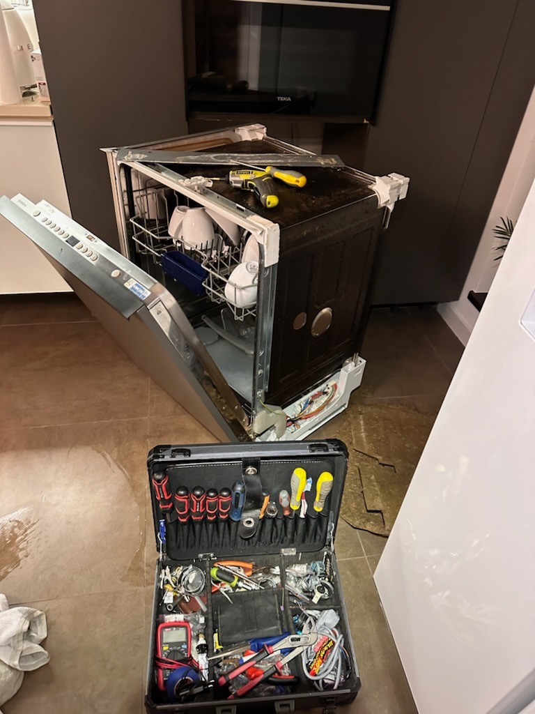 Uszkodzona zmywarka TEKA z otwartymi drzwiami, narzędzia na górnej obudowie i walizka z narzędziami na podłodze, w tle zabudowa kuchenna.