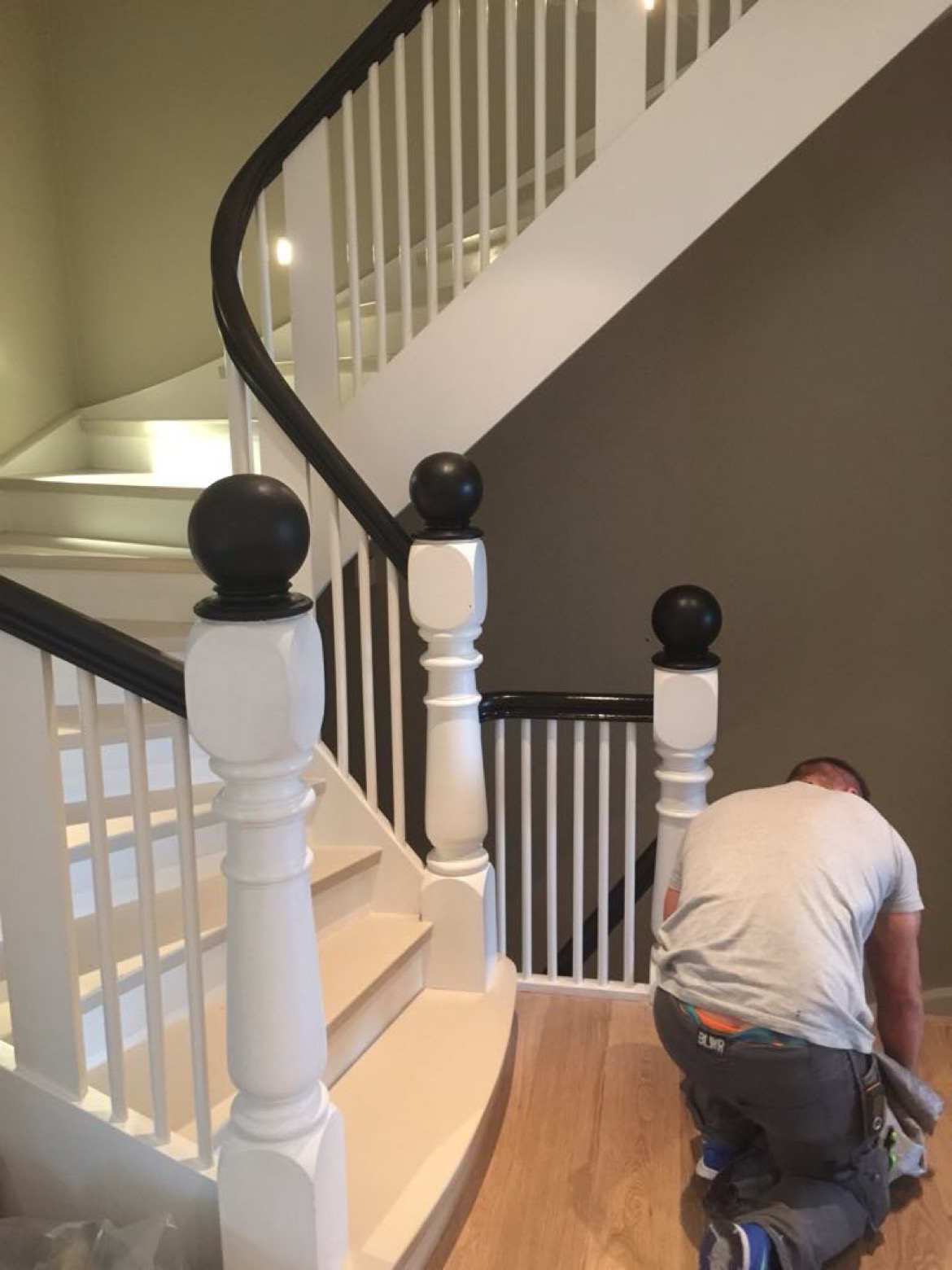 Pracownik na kolanach czyści drewnianą podłogę po malowaniu białych schodów z czarną balustradą w jasnym wnętrzu.